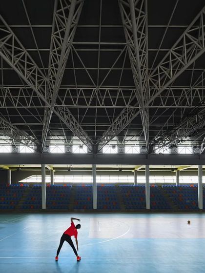 A lone athlete stretches on the vast court of the sports arena, highlighting the scale of the space. The clerestory windows along the upper level allow diffused natural light to fill the hall, reducing the need for artificial lighting during the day.