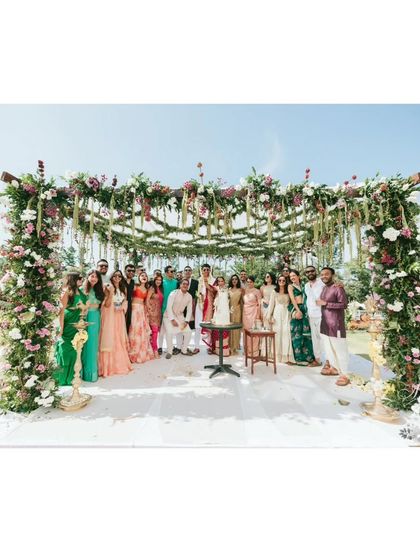 A beautiful moment as the wedding party gathers for a photo under a lush floral arch. The mandap is decorated with a canopy of greenery, hanging amaranthus, and colorful flowers.