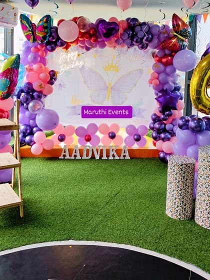 A beautiful butterfly-themed party with a purple and pink color scheme. The setup features a large backdrop, butterfly foil balloons, and a green turf floor.