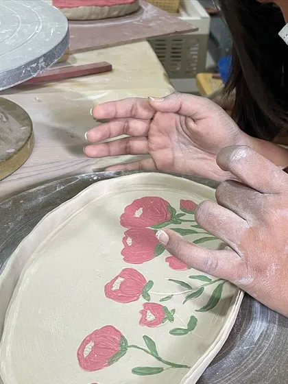 A student carefully paints delicate pink flowers onto their platter, a design that will become permanent and vibrant after glazing and firing.