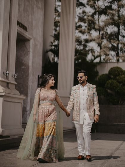 A full-length shot of the couple walking together, showcasing their beautifully detailed outfits. The modern architectural background provides a clean, sophisticated canvas for their portrait.