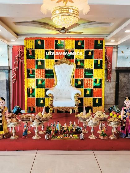 A baby shower stage in a hall with a colorful marigold grid backdrop. The setup is completed with a white throne chair and a full arrangement of traditional dolls and plates.