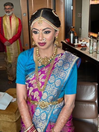 This bride is ready for her Muhurtham. Her makeup features a classic winged liner and a bold lip, perfectly complementing her bright blue and pink silk saree and traditional gold jewellery.