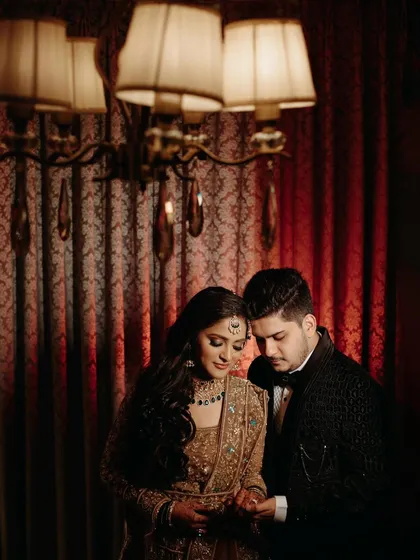 A quiet, intimate moment captured under the soft light of a chandelier. The couple looks down at the bride's hands, creating a sense of shared focus and connection.