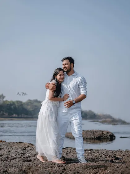 A sweet and simple embrace on the rocky shore. The couple is dressed in white, creating a clean and classic look against the natural backdrop of the river.