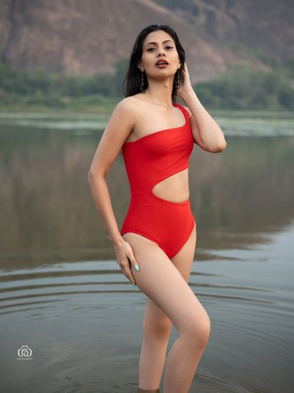 A full-body shot of a model in a red one-piece swimsuit, standing in the water. The hills in the background add a sense of scale and grandeur to this outdoor fashion shot.
