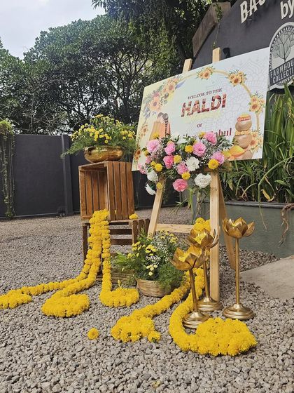 This welcome area for a Haldi ceremony is both rustic and beautiful. A personalized sign on an easel is surrounded by marigold garlands, flower arrangements, and traditional props.