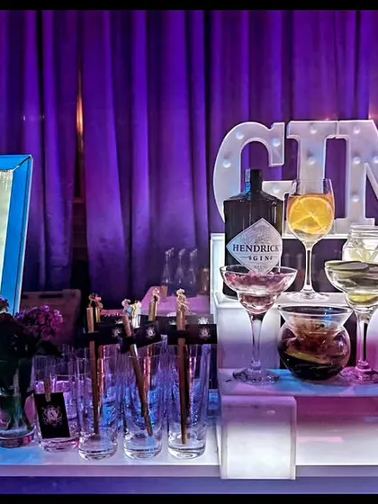 A stunning Gin Bar setup featuring a glowing menu, marquee lights, and a curated selection of gins and garnishes. The purple uplighting creates a moody and sophisticated atmosphere perfect for an evening event.