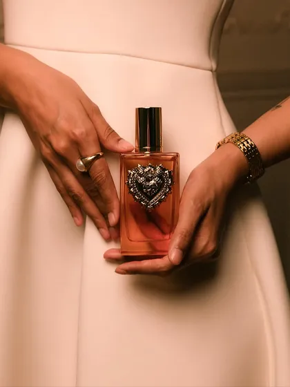 A detail shot focusing on the intricate Sacred Heart design of the D&G Devotion bottle, held against a classic white dress.