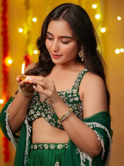 A model in a green lehenga carefully lighting a traditional diya, with a warm, glowing background of festive lights.
