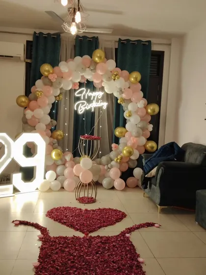 A beautiful and elegant living room setup for a 29th birthday party. This features a large circular balloon arch in pink, white, and gold, a lit-up '29' marquee number, and a path of rose petals leading to the celebration area.