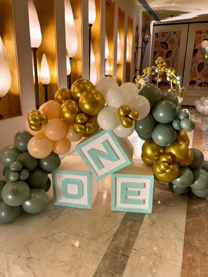 Light up "ONE" blocks surrounded by a garland of sage green, gold, and peach balloons, used as a decorative accent for the twins' first birthday party.