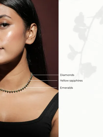 The Leafline Necklace worn by a model, showcasing its delicate sparkle and how it sits beautifully on the collarbone. The gems are labeled to highlight the combination of diamonds, yellow sapphires, and emeralds.