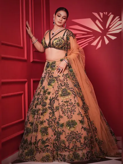 A dynamic pose against a bold red background. The contrast makes the green and peach embroidery of the lehenga pop, creating a visually striking image.