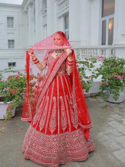 The bride holding her veil, showcasing the full length of the red lehenga against a beautiful white building. The contrast makes the outfit pop.