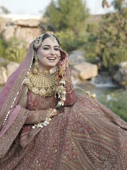 A moment of pure bridal bliss. This bride, dressed in a dusty rose lehenga with heavy gold jewelry, shares a laugh, showcasing her radiant makeup in a beautiful outdoor setting.