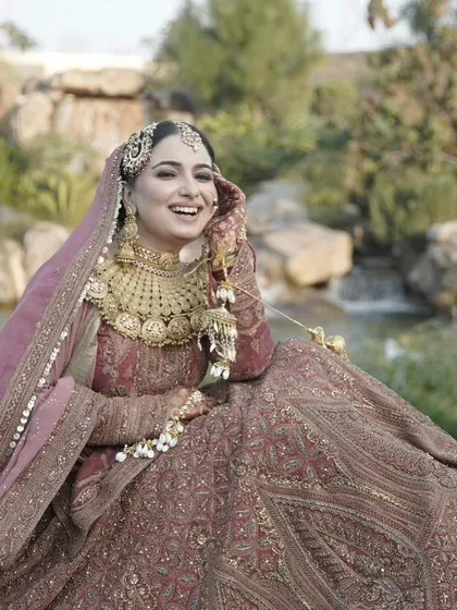 A moment of pure bridal bliss. This bride, dressed in a dusty rose lehenga with heavy gold jewelry, shares a laugh, showcasing her radiant makeup in a beautiful outdoor setting.