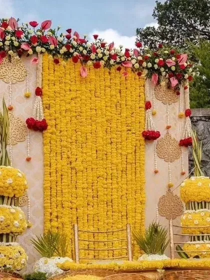 A traditional Haldi or Pellikuthuru ceremony backdrop. The wall of yellow marigolds is accented with red flowers and traditional motifs.