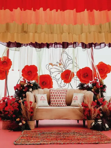 The main seating for the couple at a vibrant Mehendi, featuring a backdrop of giant red paper flowers and a comfortable sofa with patterned cushions.