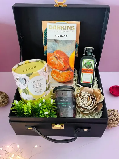 A refined Diwali hamper in a black suitcase box. It contains a "Lost in Garden" scented candle, orange chocolate, coffee scrub, and a miniature liqueur.