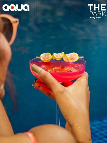 A close-up of a beautifully garnished pink cocktail held against the backdrop of the pool. We focus on craft and presentation, ensuring every drink is an experience.