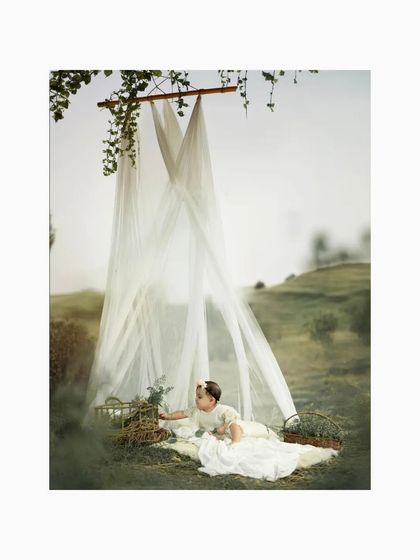 A baby girl lies on a soft blanket under a dreamy canopy in a green, rolling field. This ethereal and gentle setup creates a magical, fairy-tale-like setting for a first birthday photoshoot.