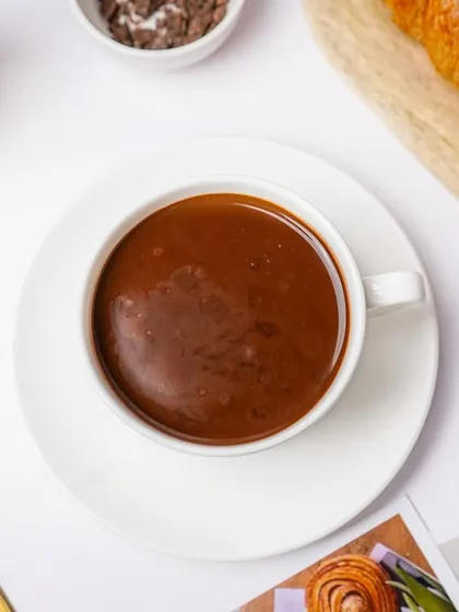A top-down view of a cup of thick hot chocolate, styled with a croissant and a golden spoon. This flatlay composition creates a cozy and inviting cafe vibe.