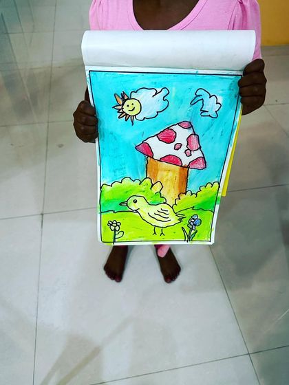 A proud student showing off her colourful drawing of a bird next to a mushroom. The bright colours and cheerful sun make this a very happy picture.
