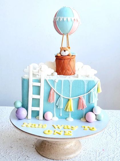 A "Halfway to One" cake for a 6-month birthday. The design features a hot air balloon, a ladder to the clouds, and a cute teddy bear topper.