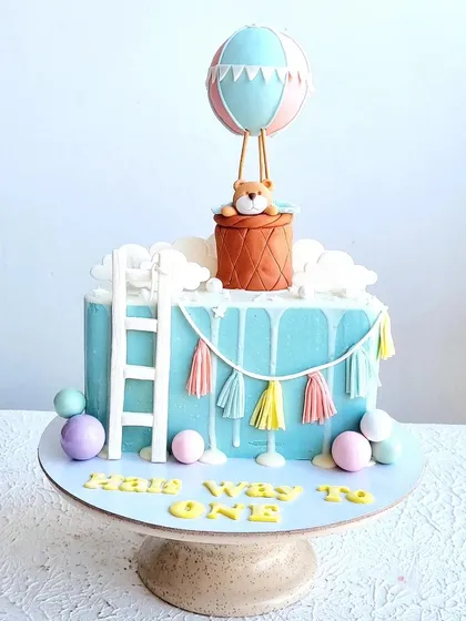 A "Halfway to One" cake for a 6-month birthday. The design features a hot air balloon, a ladder to the clouds, and a cute teddy bear topper.