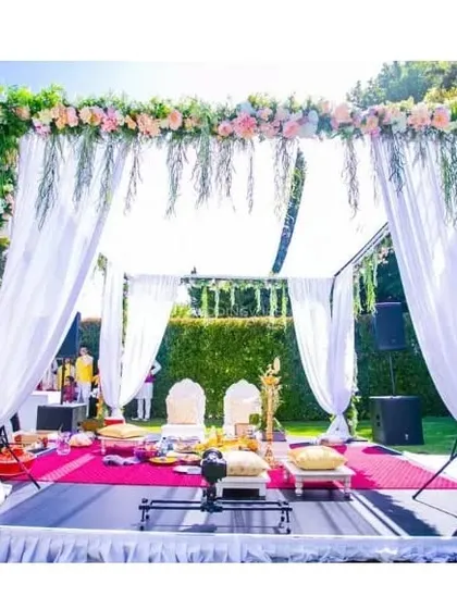 A beautiful outdoor wedding mandap with a lush green backdrop. The structure is decorated with a mix of flowers and greenery, with white drapes adding a touch of elegance.