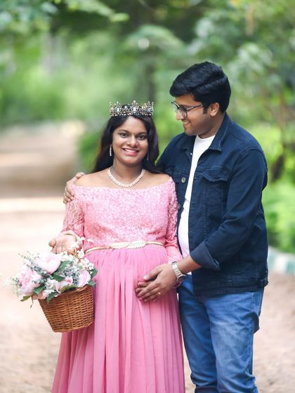 A happy couple portrait taken on a park path. The dad-to-be has his arm around his partner, who is wearing a lovely pink dress and a crown.