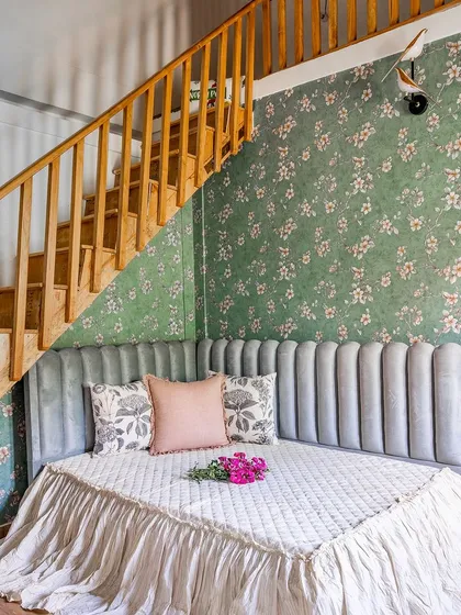 A cozy reading nook for a child, designed under a staircase with a custom-cushioned daybed, plush headboard, and charming floral wallpaper.