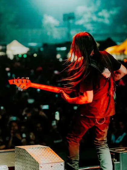 A dynamic photo of a long-haired bassist on stage, captured mid-performance in front of a massive festival crowd.