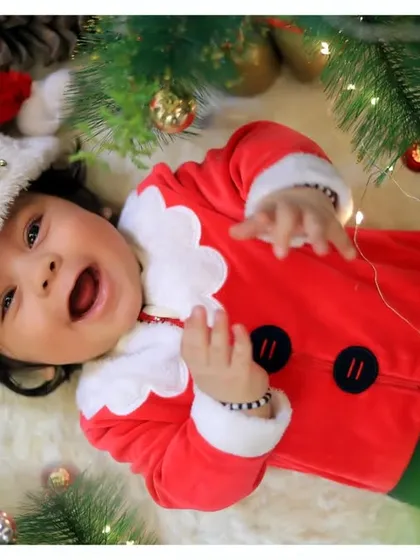 A joyful baby celebrating his first Christmas, lying among the pine branches and festive lights in a cute Santa outfit.