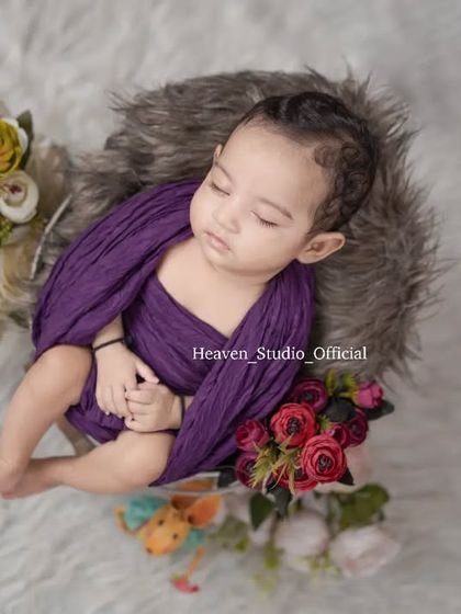 A newborn boy rests peacefully in a fur-lined basket, wrapped in a rich purple swaddle. The surrounding flowers add a touch of color and softness to the composition.