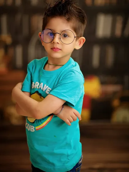 A close-up portrait of a boy with glasses and his arms crossed, looking very smart and studious against a library backdrop.