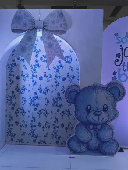 A close-up of a baby boy shower backdrop. This setup features a large gift box design with a floral pattern, a cute teddy bear cutout, and the phrase "so much joy in this little boy".