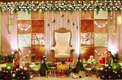 A beautiful baby shower stage with a backdrop of jasmine and marigold panels. The setup is adorned with peacock figures, traditional dolls, and a full display of offerings, creating a scene of prosperity.