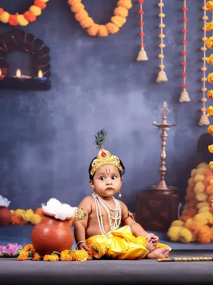 A baby boy dressed as Krishna, sitting in a festive setup decorated with marigold garlands and earthen pots.