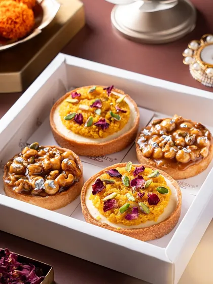 A close-up of four exquisite mini tarts in a white box, two topped with motichoor and pistachio, and two with a nutty caramel glaze. These fusion desserts are a perfect example of how I blend traditional Indian flavors with classic pastry techniques.