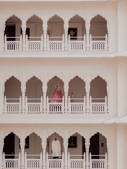 A breathtaking wide shot showing the bride and groom on separate balconies of a palace, a visual metaphor for two lives about to join. This architectural portrait highlights the grandeur of a destination wedding venue.