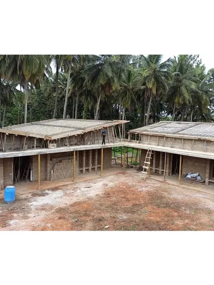 An aerial view during the construction phase, showing the two building blocks arranged around the central courtyard. The structure is set against a backdrop of coconut groves, highlighting its integration into the local landscape.