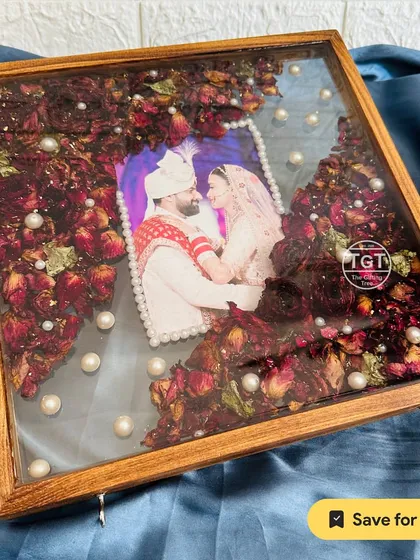A 12-inch square teakwood frame with preserved red rose petals and a hanging nameplate featuring a wedding hashtag and date.