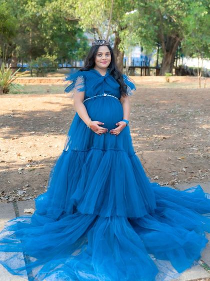 A full-length shot of a mom-to-be in a royal blue gown and tiara, looking like a queen in a park setting. The gown's long train is spread out beautifully.