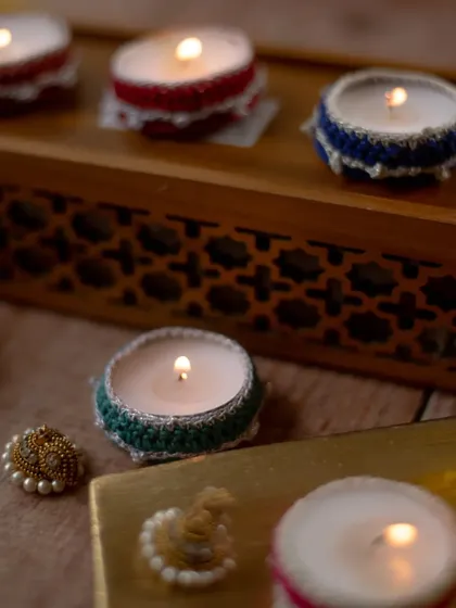 Our crochet tea light holders in various colors, shown lit in a cozy setting. These small 'diya jackets' are perfect for scattering around the house to create a warm, festive glow.