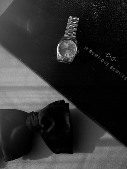 The groom's accessories, a classic watch and a bow tie. This flat-lay style shot, in moody black and white, captures the small details that complete the wedding day look.