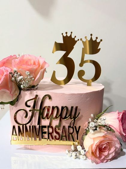 A one-kg anniversary cake in a soft pink, decorated with fresh roses, baby's breath, and a golden "Happy Anniversary" topper.
