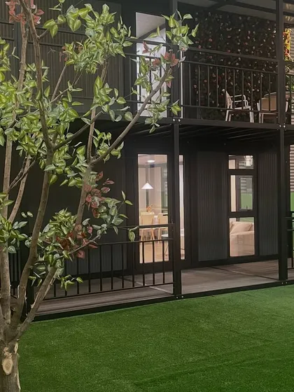 A side view of the two-story container home, highlighting the wrap-around porch and balcony. The black steel frame provides a strong visual contrast to the surrounding greenery.