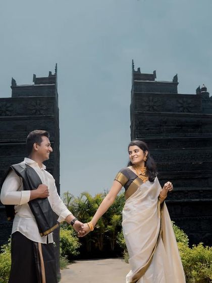 A couple in traditional South Indian attire walking hand-in-hand, framed by two impressive black stone structures, creating a visually striking and culturally rich photograph.